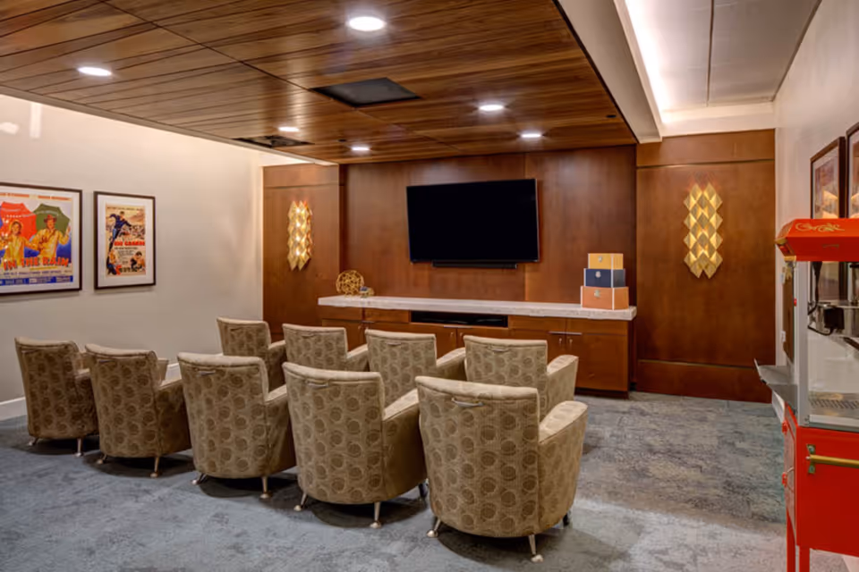 A cozy media room with two rows of patterned armchairs facing a large flat-screen TV mounted on a wooden paneled wall. The room has a wooden ceiling with recessed lighting, framed vintage movie posters on one wall, and a red popcorn machine on the right side.