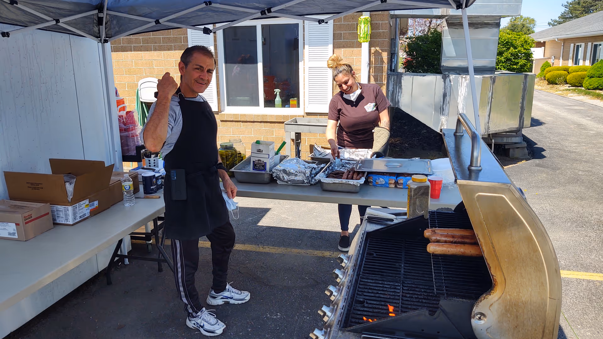 Two people cooking outdoors under a canopy tent. One person is standing near a table with food items and the other is grilling sausages on a large outdoor grill. The setting appears to be a parking lot next to a building.