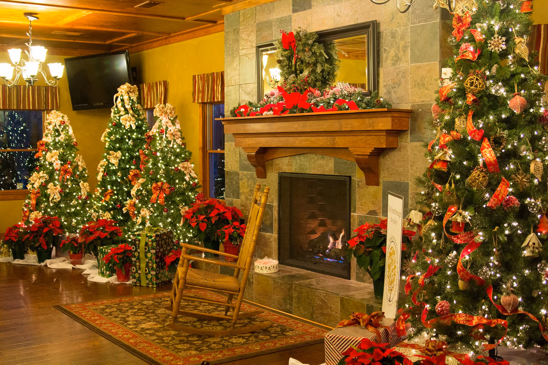 A cozy living room decorated for Christmas with multiple decorated Christmas trees, poinsettia plants, a stone fireplace with a wooden mantle adorned with holiday greenery and red bows, a rocking chair on a patterned rug, and wrapped presents under the trees.