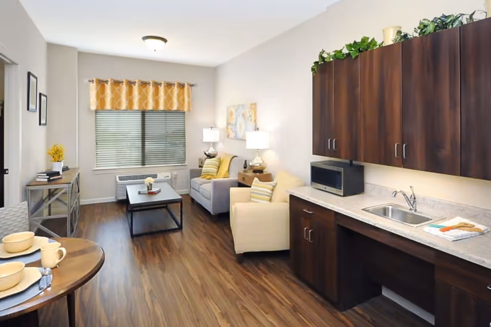 Bright combined living area and kitchenette with a sofa, armchair, coffee table, dining nook, and dark wood cabinets.