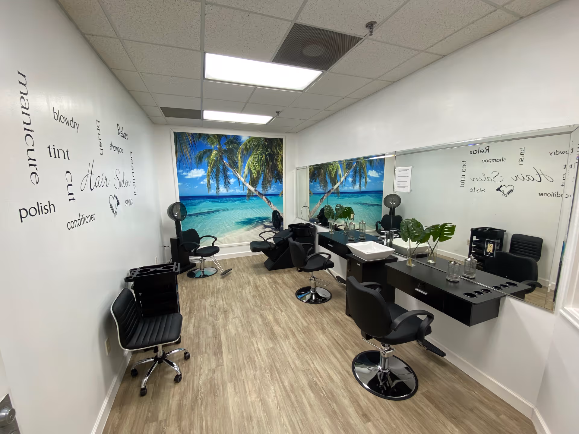 Interior of a hair salon room with black salon chairs, a large mirror on the right wall, and a tropical beach mural on the back wall. The left wall has words related to hair care such as manicure, polish, tint, cut, conditioner, blowdry, shampoo, relax, and style. The floor is wood-patterned, and the ceiling has fluorescent lighting.
