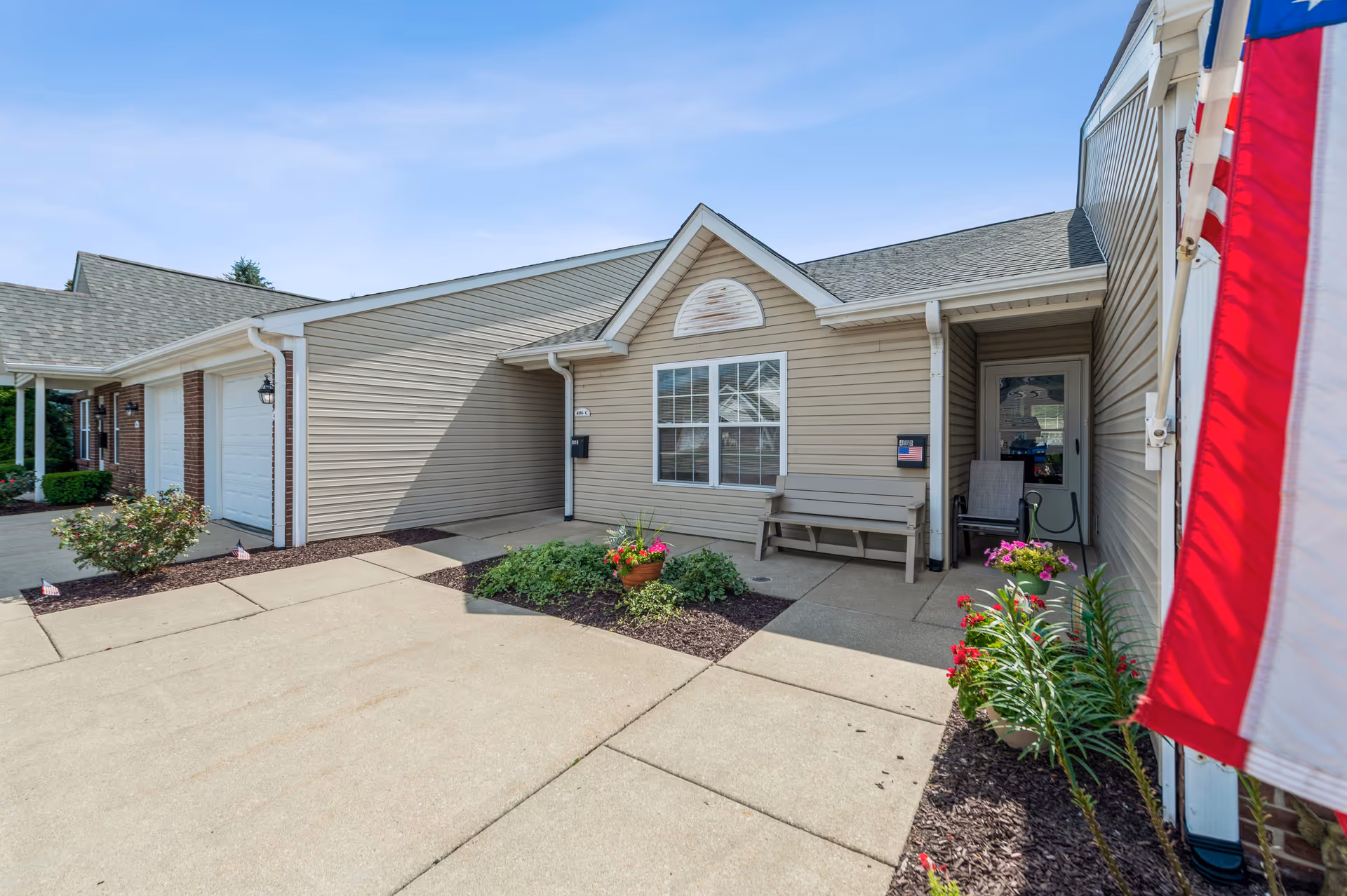 Exterior view of a residential-style building with beige siding and a gray shingled roof. There is a concrete driveway and walkway leading to a small porch area with a bench and potted flowers. An American flag is visible on the right side of the image. The sky is clear and blue.