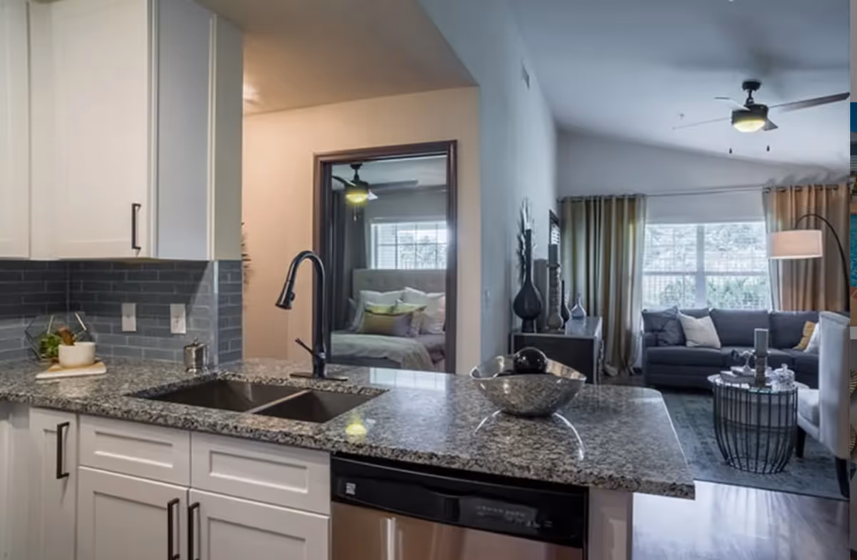 Modern kitchen with granite countertop, double sink, and black faucet. White cabinets with black handles and a gray tile backsplash. In the background, a bedroom with a bed and ceiling fan is visible through a doorway, and a living room with a gray sofa, armchair, coffee table, floor lamp, and large window with curtains is seen.