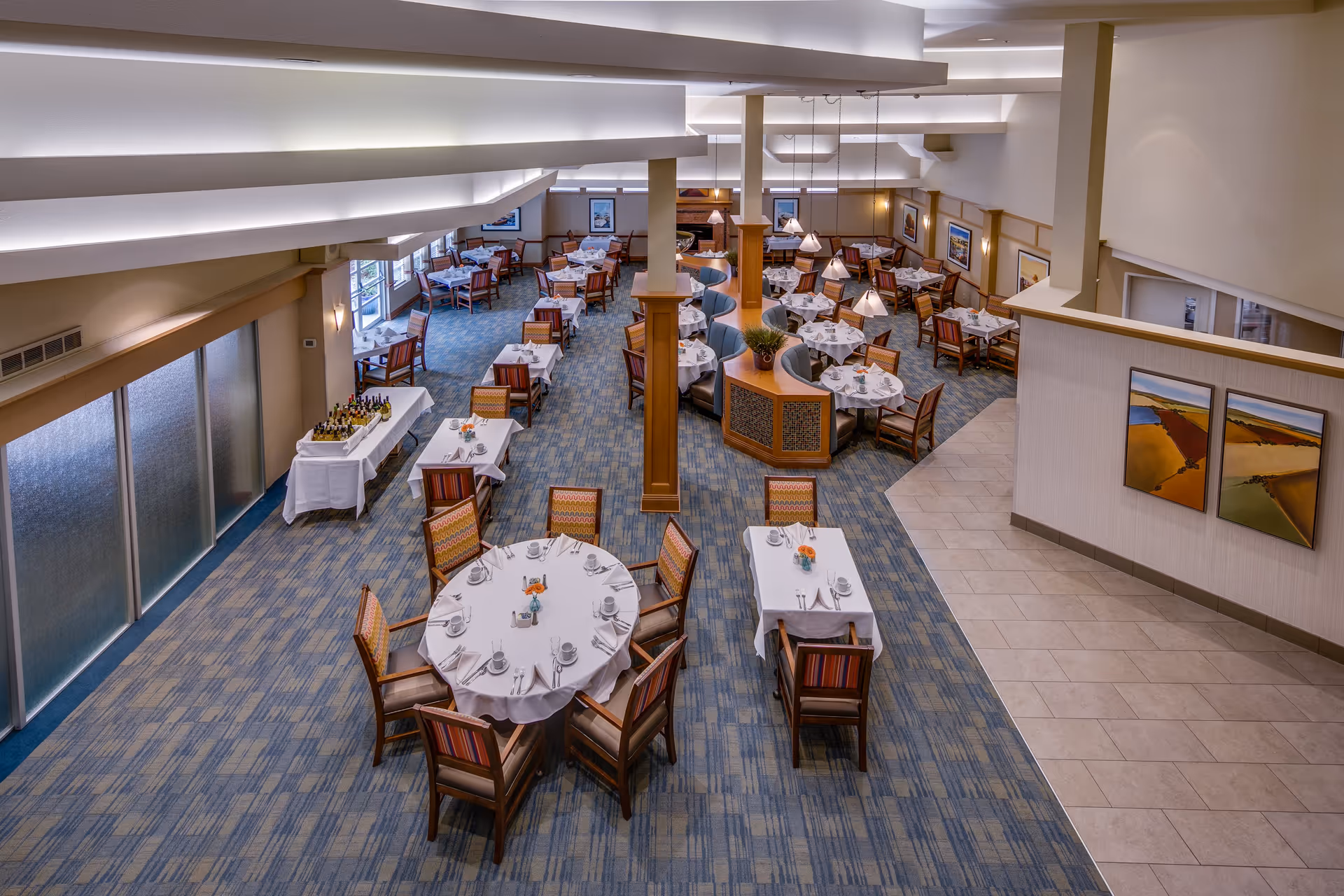 A spacious dining room with multiple tables covered in white tablecloths, each set with cups, saucers, and silverware. The room features carpeted flooring with a blue pattern, wooden chairs with colorful upholstery, and soft overhead lighting. There are paintings on the walls and a table with bottles and glasses near frosted glass doors on the left side.