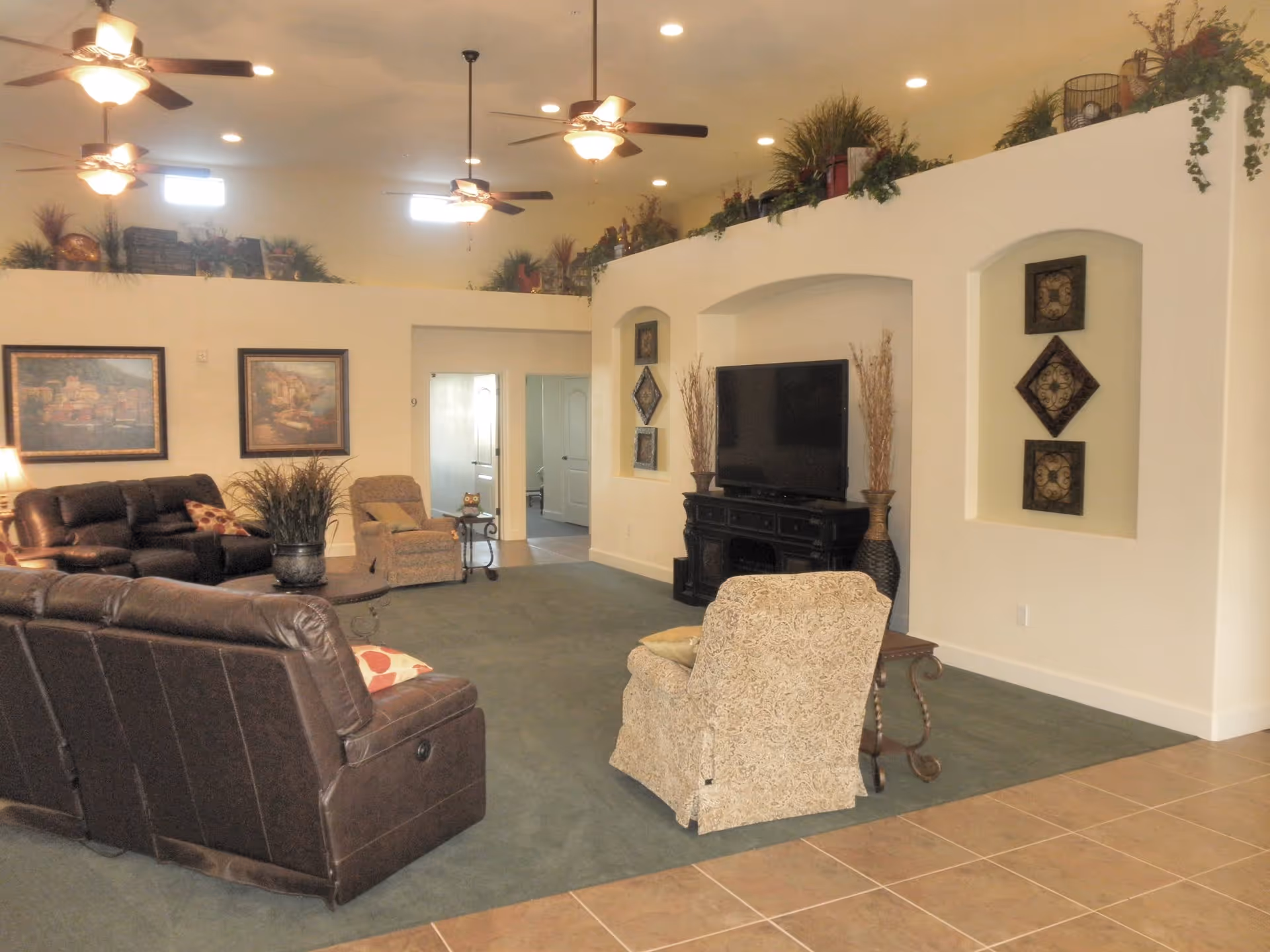 A spacious living room in an assisted living facility featuring multiple ceiling fans with lights, a large flat-screen TV on a black stand, leather and fabric armchairs and sofas arranged around a central coffee table, framed artwork on the walls, and decorative plants placed on a ledge near the ceiling.