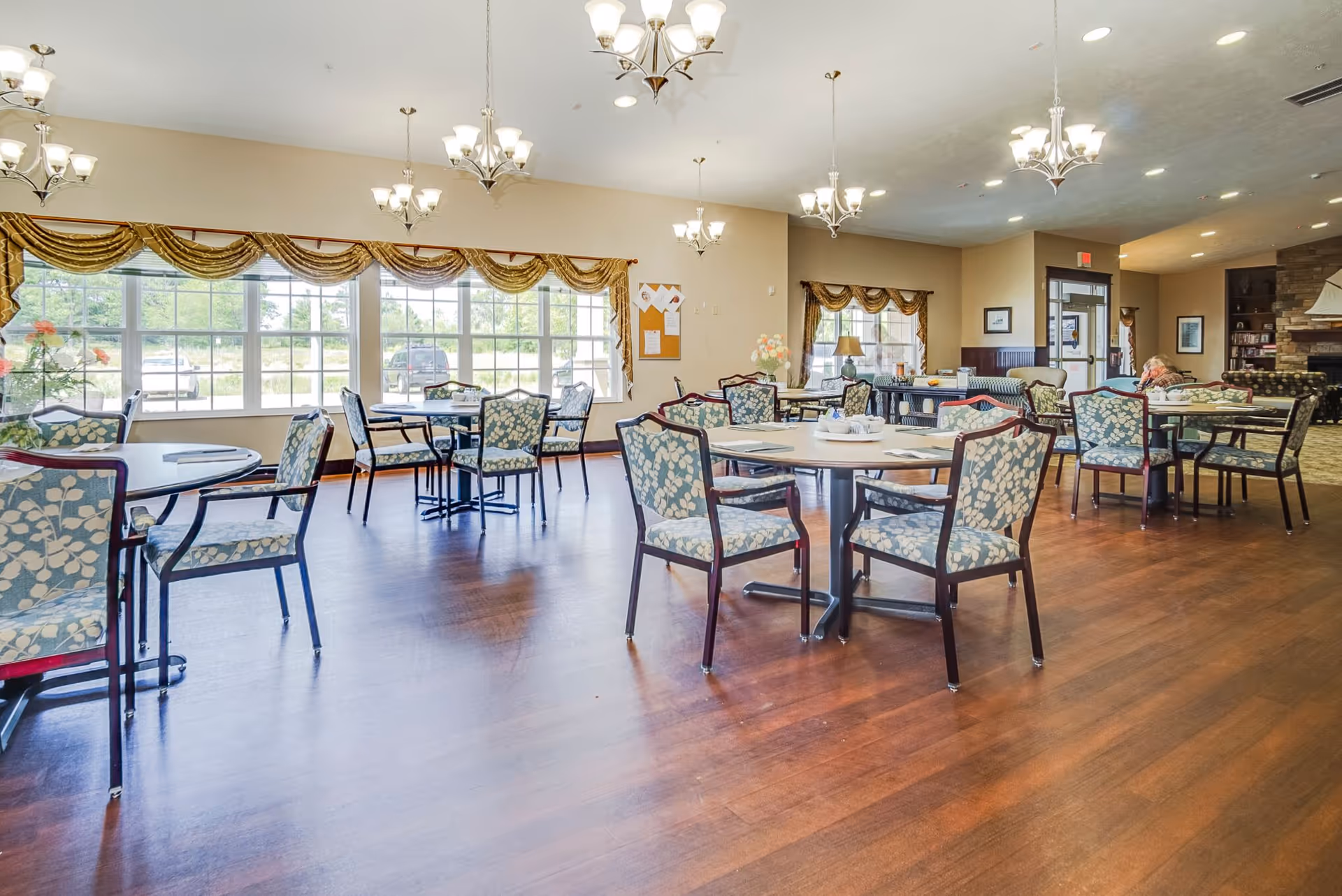A spacious dining room with multiple round tables and chairs upholstered in a blue and white floral pattern. Large windows with gold valances let in natural light, and several chandeliers hang from the ceiling. The room has wooden flooring and a cozy seating area with a fireplace in the background.