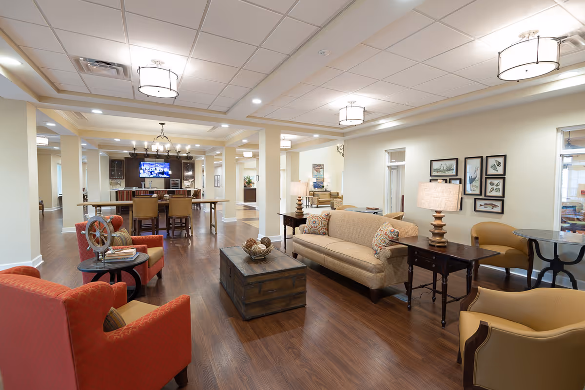 A spacious and well-lit living room area in a senior living facility featuring a beige sofa with patterned cushions, two red armchairs, two tan armchairs, a wooden coffee table with decorative items, side tables with lamps, and framed artwork on the walls. In the background, there is a dining area with a long wooden table, chairs, and a mounted flat-screen TV.