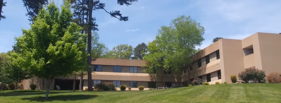 Exterior view of a two-story beige building surrounded by green grass, trees, and shrubs under a clear blue sky.