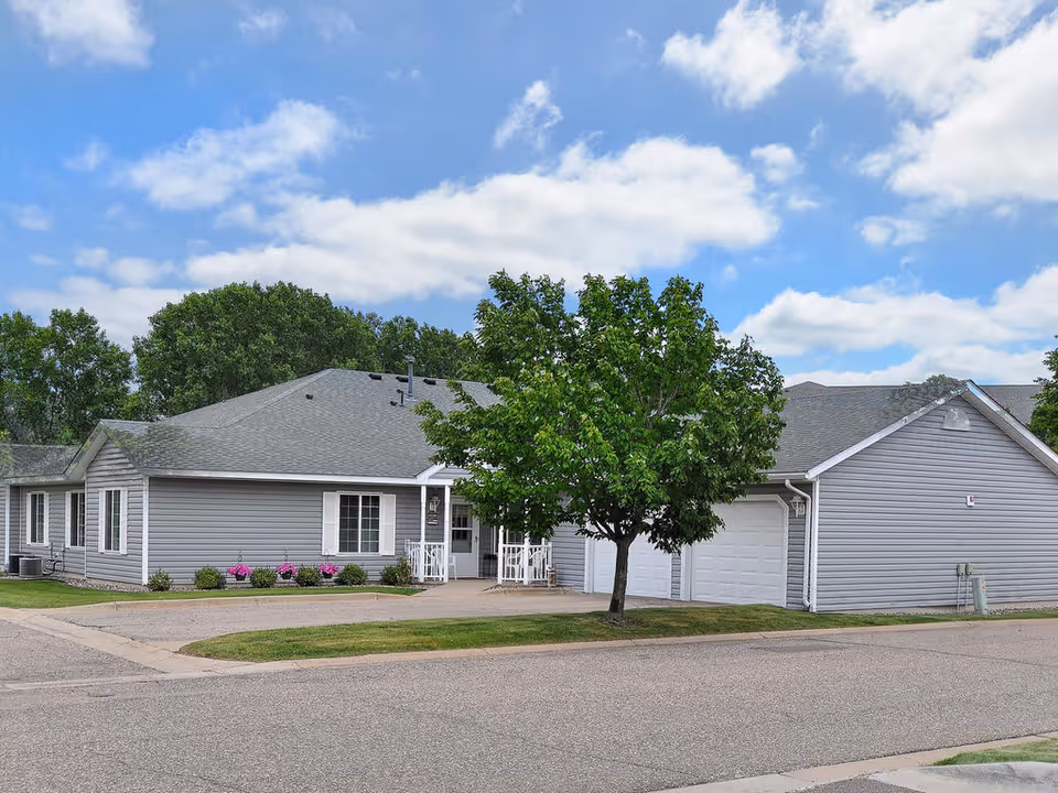 Exterior view of a single-story gray building with white trim, a small porch with white railings, a tree in front, and a garage door on the right side under a partly cloudy blue sky.