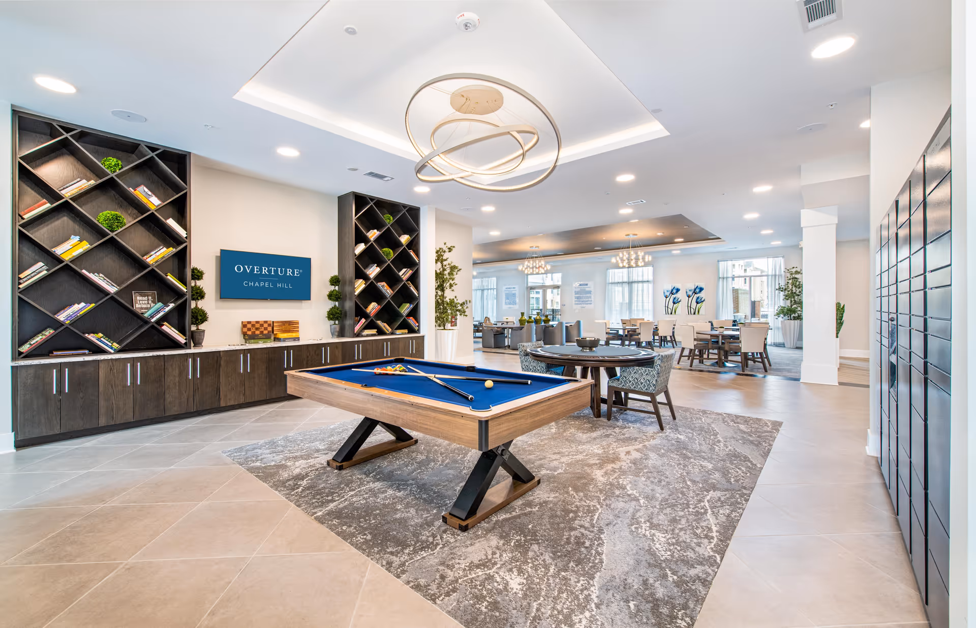 Bright communal lounge with a pool table, seating areas, bookshelves, and dining tables in a modern open interior.