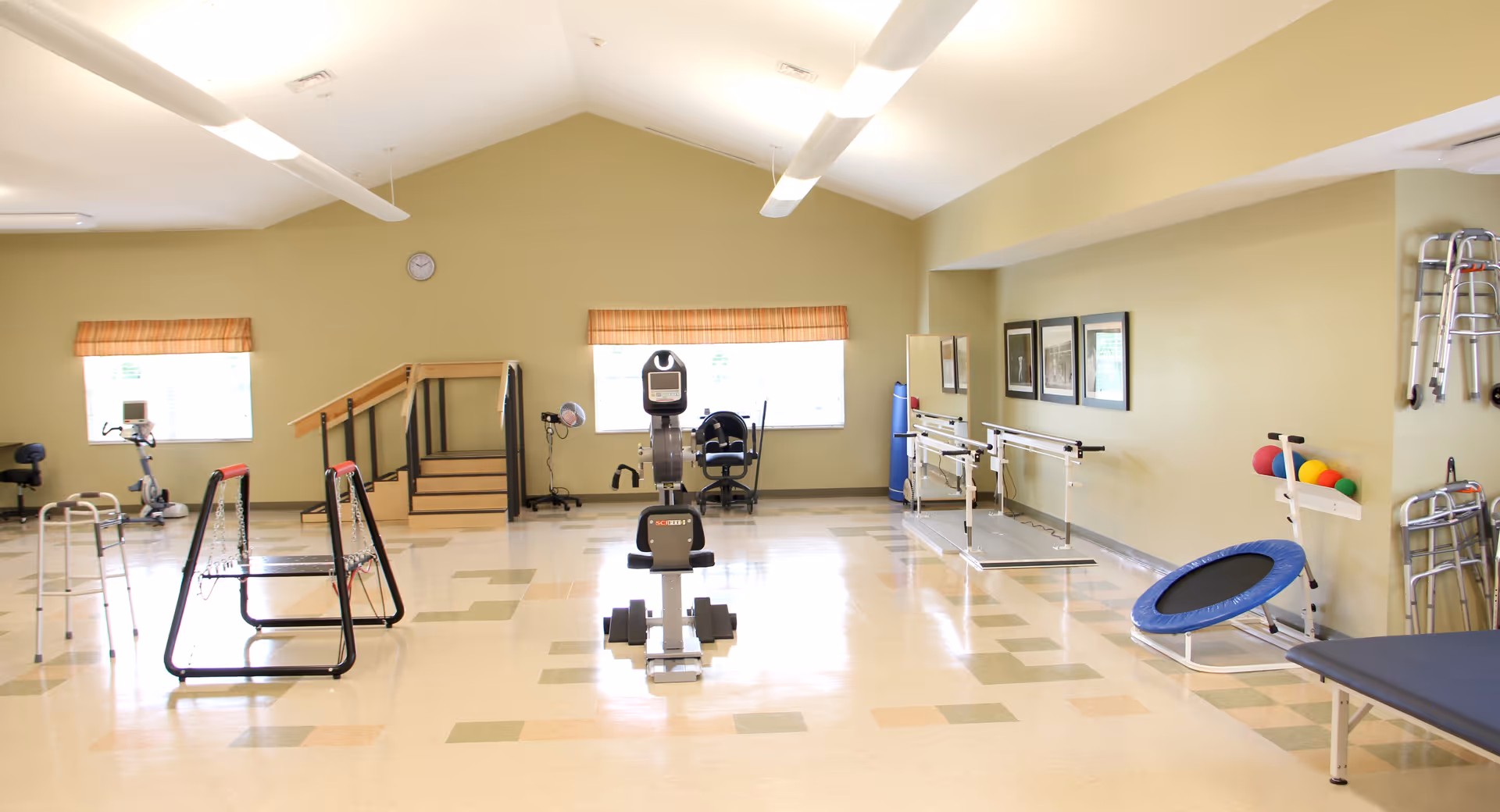 A spacious rehabilitation or physical therapy room with exercise equipment including a stationary bike, parallel bars, a small trampoline, walkers, and therapy balls. The room has light green walls, two windows with orange blinds, and a clock on the wall. There are framed pictures on one wall and a treatment table on the right side.