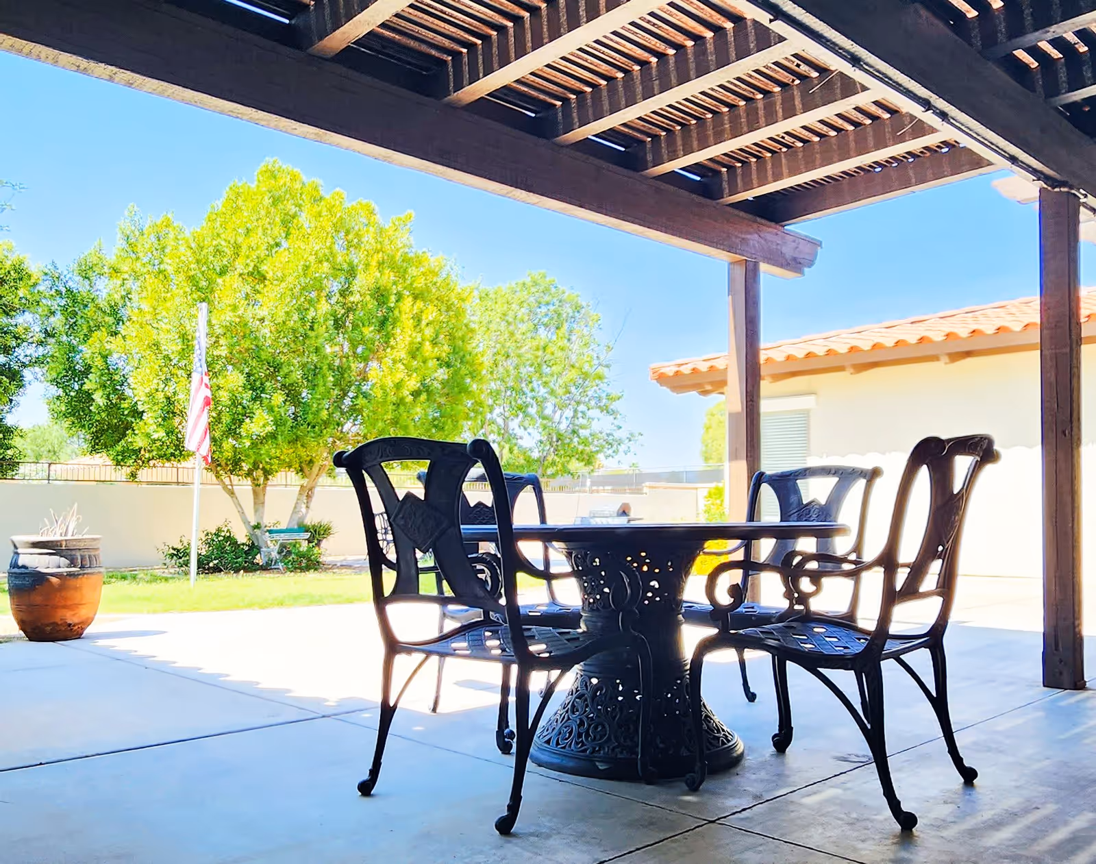 Outdoor patio area with a round metal table and four matching chairs under a wooden pergola. In the background, there is a green tree, an American flag on a flagpole, a large planter, and a building with a tiled roof.