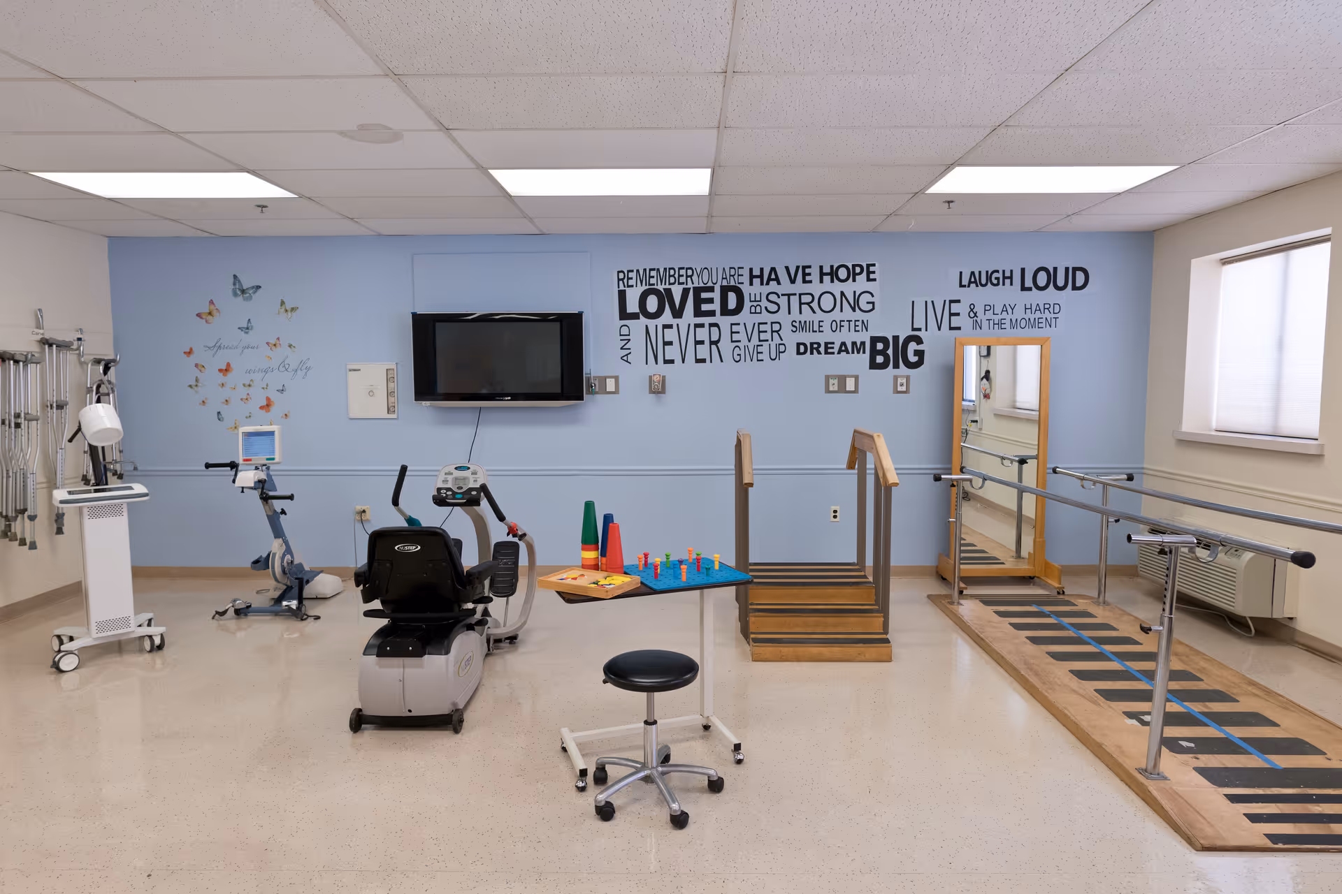 A rehabilitation therapy room with exercise equipment including stationary bikes, a step platform with handrails, parallel bars for walking practice, a stool, and a table with colorful therapy tools. The walls are light blue with motivational quotes and butterfly decals. A flat-screen TV is mounted on the wall, and there is a large mirror near the parallel bars. The room is well-lit with ceiling lights and has a window with blinds.