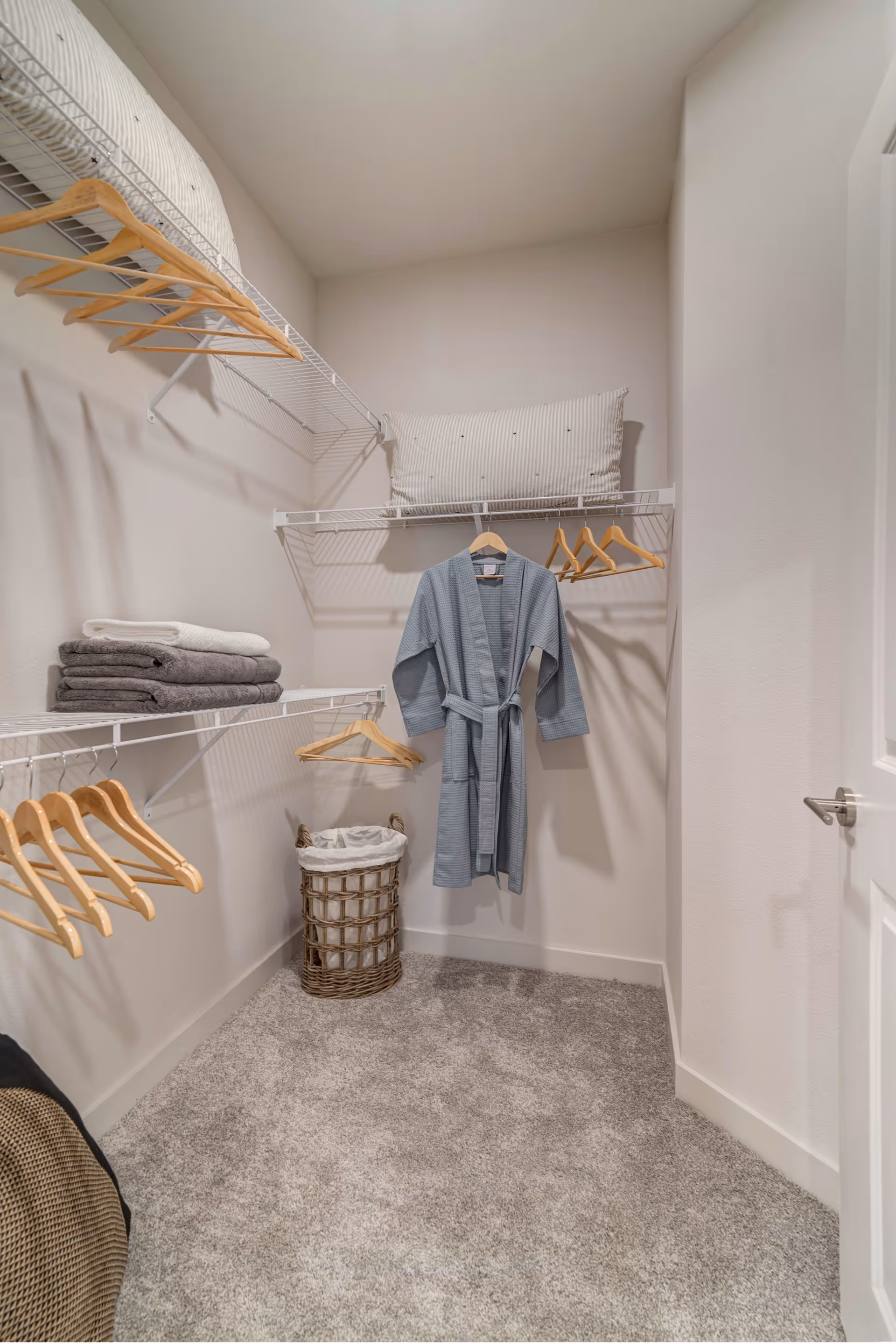 A walk-in closet with beige walls and carpeted floor. The closet has white wire shelving on three walls, holding wooden hangers, folded towels, a striped pillow, and a blue robe hanging on the back wall. A woven laundry basket with a white liner is on the floor beneath the robe.