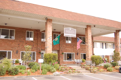 Exterior view of a two-story brick building with a covered entrance supported by columns. There are two flags displayed, one is the American flag and the other is a green flag. The entrance is surrounded by well-maintained landscaping with bushes and flowers. A sign hanging from the ceiling reads 'Emeritus Senior Living'.