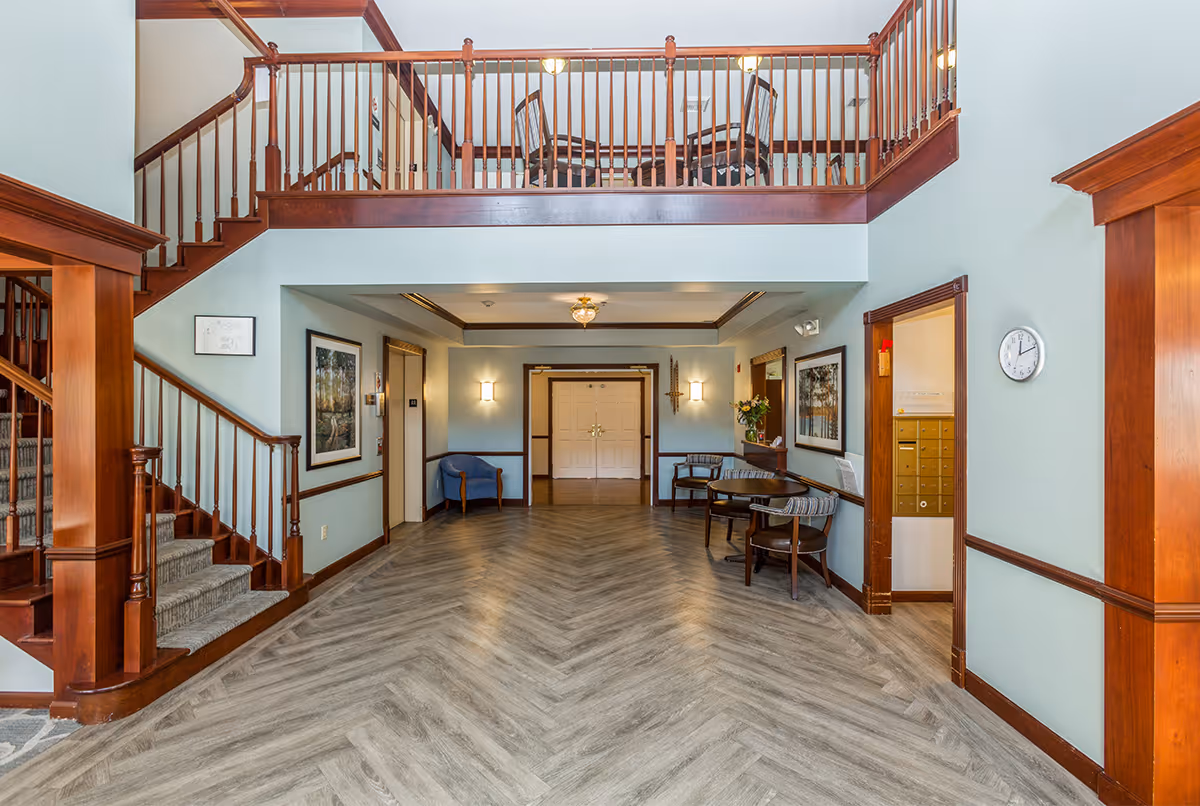 Interior view of a senior living facility lobby with wooden staircase on the left, a balcony with wooden railing above, light blue walls, framed artwork, a clock on the right wall, and several chairs and small tables arranged along the walls.