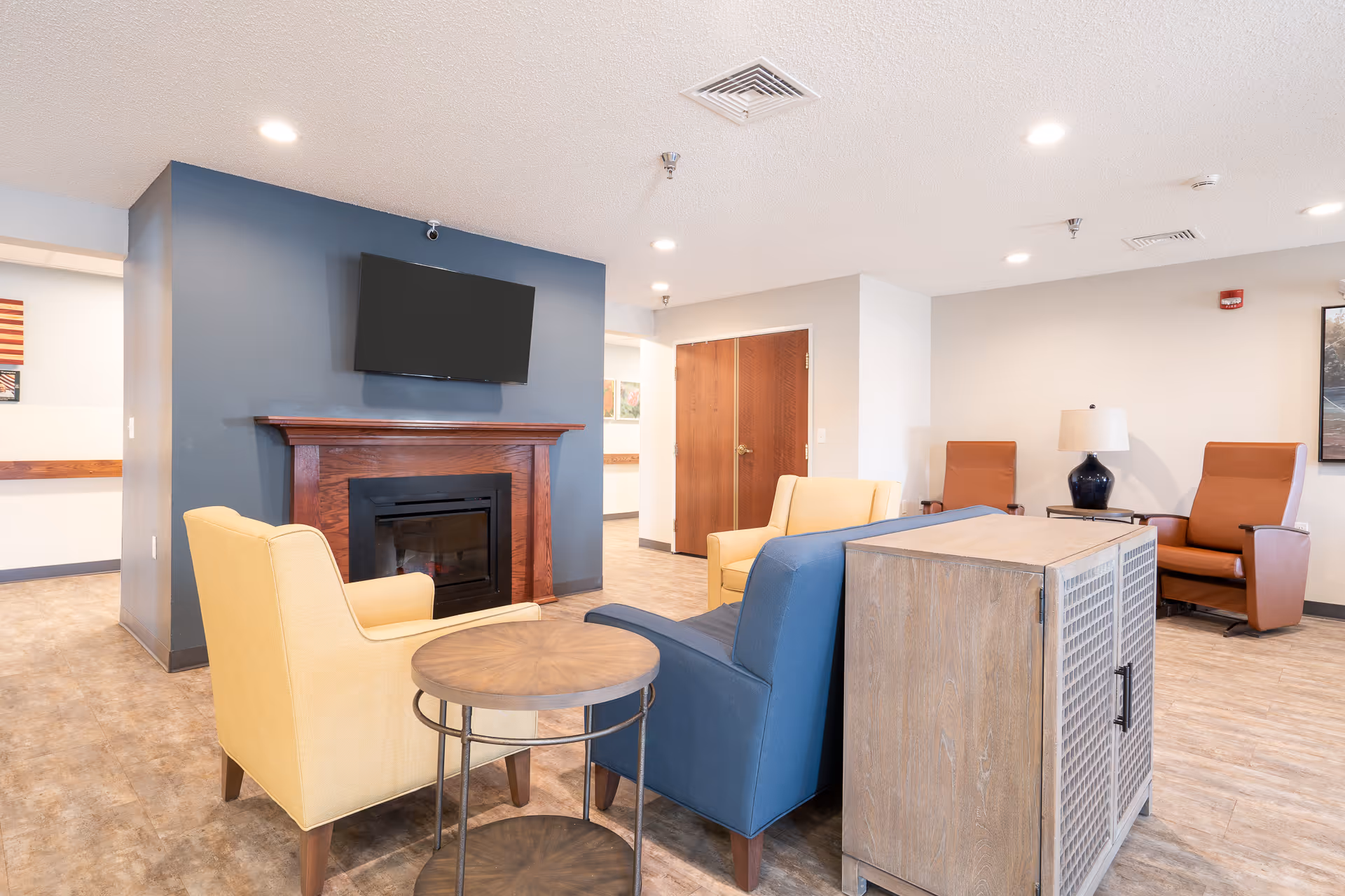 A bright and spacious living room area in a senior living facility featuring a blue accent wall with a mounted flat-screen TV above a wooden fireplace. The room includes comfortable seating with two yellow armchairs, a blue armchair, and two brown recliners. There is a round wooden side table and a wooden cabinet, with a lamp on a small table near the recliners. The floor is covered with light-colored wood-style flooring, and the ceiling has recessed lighting.