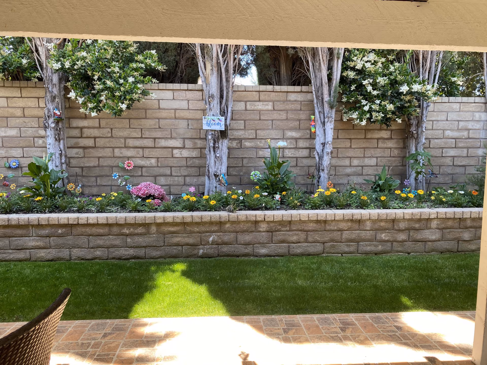 View of a small outdoor garden area with a raised flower bed containing various colorful flowers and plants. Behind the flower bed is a brick wall with several trees growing in front of it. There are decorative flower and butterfly ornaments placed among the plants. The foreground shows a tiled patio and a patch of green grass.