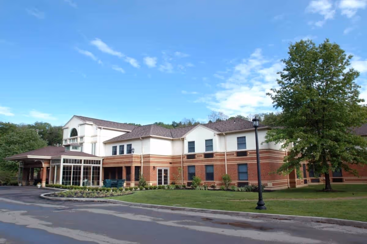 Exterior view of a two-story senior living facility building with a combination of brick and light-colored siding, surrounded by greenery and a clear blue sky.