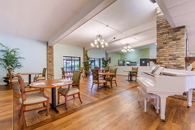 Bright dining room with round tables and chairs, a white grand piano, chandeliers, brick columns, and a serving counter.