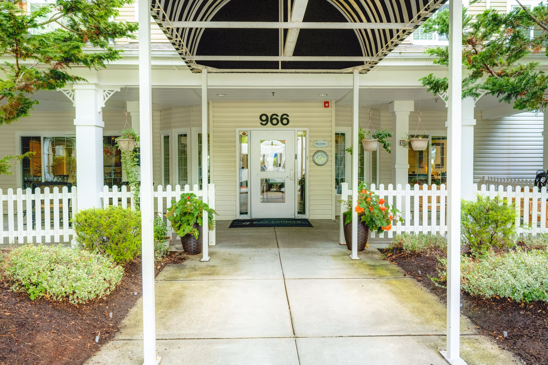 Entrance to a senior living facility with the number 966 above the door. The entrance is covered by a black and white striped awning supported by white pillars. There are potted plants with flowers on either side of the walkway and a white picket fence surrounding the area. Green shrubs and trees are visible around the entrance.
