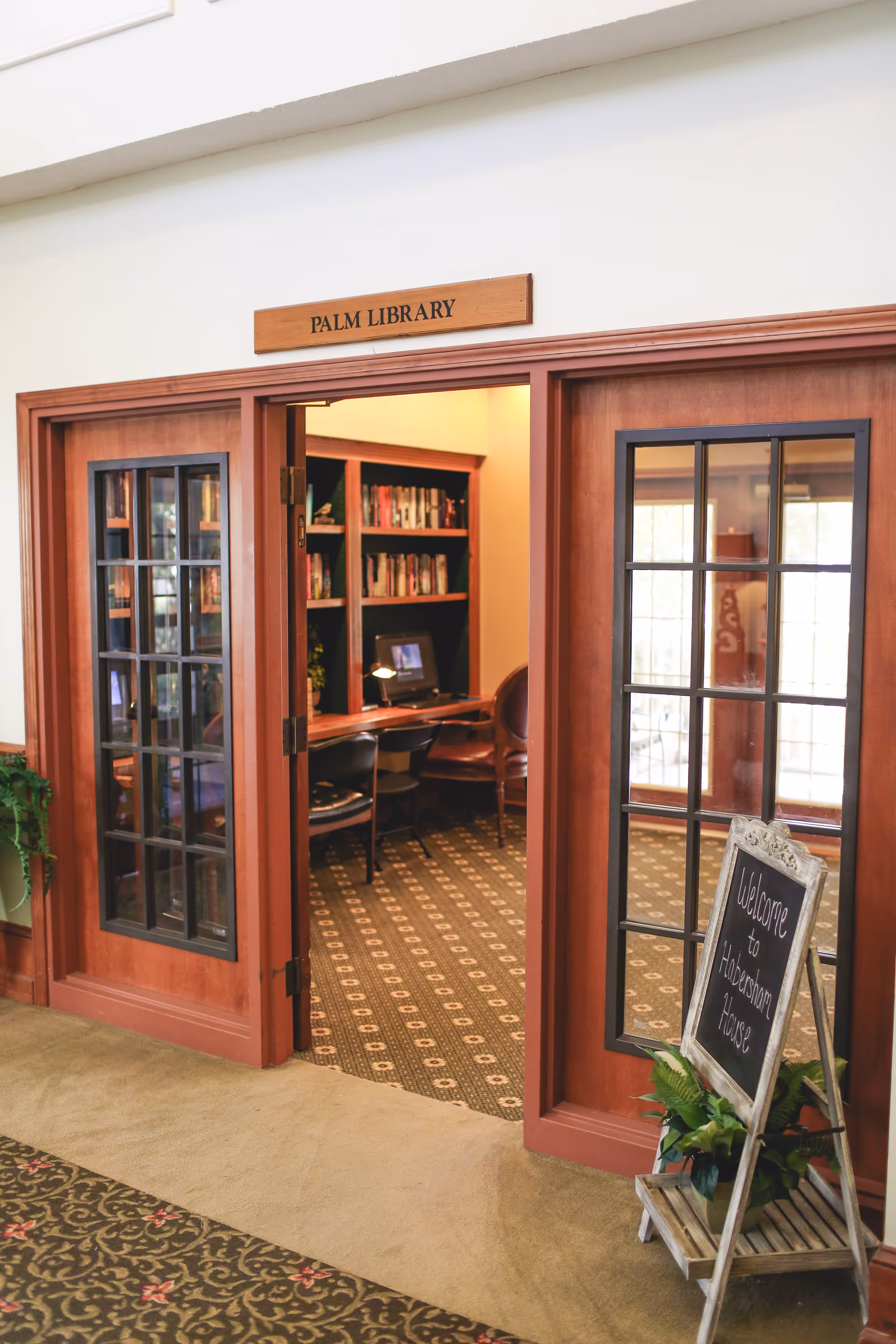 Entrance to the Palm Library with wooden double doors featuring glass panes. Inside, there are bookshelves filled with books, a desk with a computer, and chairs. A small chalkboard sign near the entrance reads 'Welcome to Habersham House' with a potted plant beneath it.