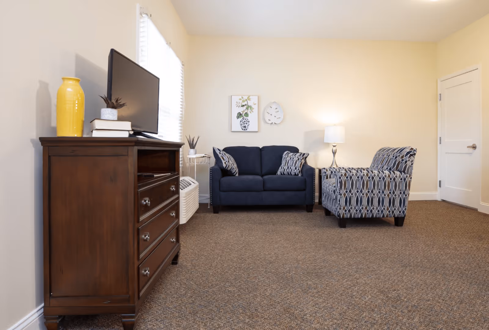 A cozy living room with a dark wooden dresser holding a flat-screen TV and decorative items, a navy blue loveseat with patterned pillows, a patterned armchair, a floor lamp, and wall art on a beige wall.