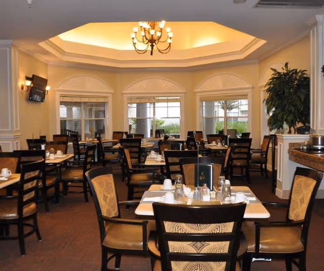 Dining room with multiple set tables and chairs, a central chandelier, and windows under a coffered ceiling.