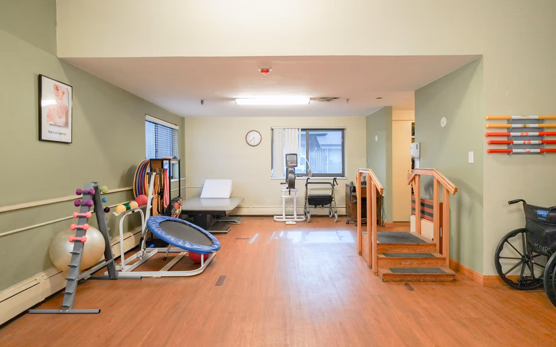 A physical therapy or rehabilitation room with exercise equipment including a small trampoline, dumbbells, exercise balls, parallel bars with wooden handrails, a stationary bike, and a walker. The room has light green walls, wooden flooring, a window, and a clock on the wall.