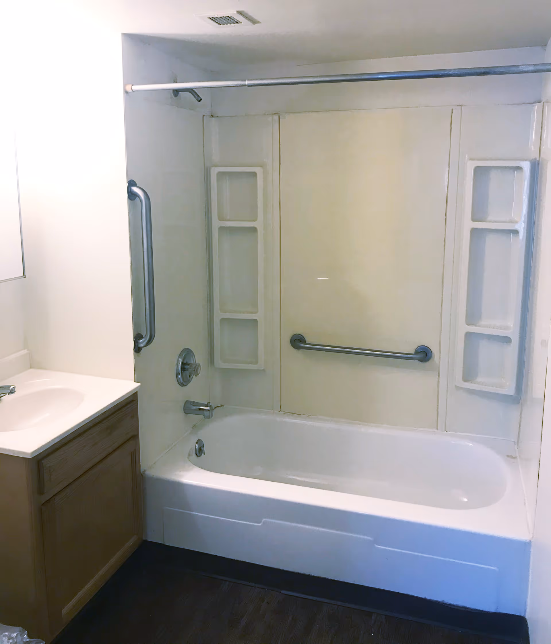 A bathroom with a white bathtub and shower combination. The bathtub has built-in shelves on both sides and a horizontal grab bar on the back wall. There is a vertical grab bar on the left wall near the faucet and shower controls. To the left of the bathtub is a wooden vanity with a white sink and faucet. The floor is dark-colored, and the walls are light-colored.