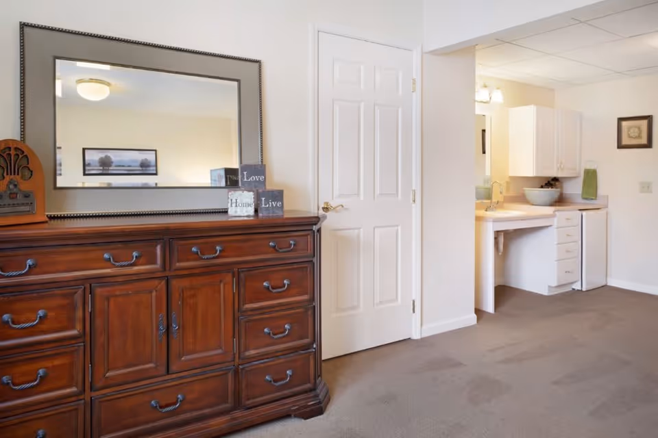 Interior view of a room featuring a large wooden dresser with multiple drawers and a large mirror above it. On the dresser are decorative blocks with the words 'Love', 'Home', and 'Live'. To the right, there is a white door and a small kitchenette area with white cabinets, a sink, a bowl, a small refrigerator, and a green towel hanging on the wall. The room has beige walls and carpeted flooring.
