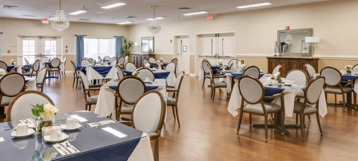 A spacious dining room in a senior living facility with multiple round tables covered with white and blue tablecloths, each set with cups, saucers, utensils, and small flower arrangements. The room has wooden flooring, beige walls, large windows with blue curtains, chandeliers, and a sideboard with a mirror and coffee setup.