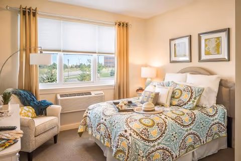 A cozy bedroom in a retirement community featuring a bed with colorful patterned bedding and multiple pillows. There is a beige armchair with a blue throw and a yellow pillow next to a floor lamp. A window with beige curtains and a white blind lets in natural light, and an air conditioning unit is installed below the window. Two framed artworks hang on the wall above the bed, and a bedside table with a lamp is visible.