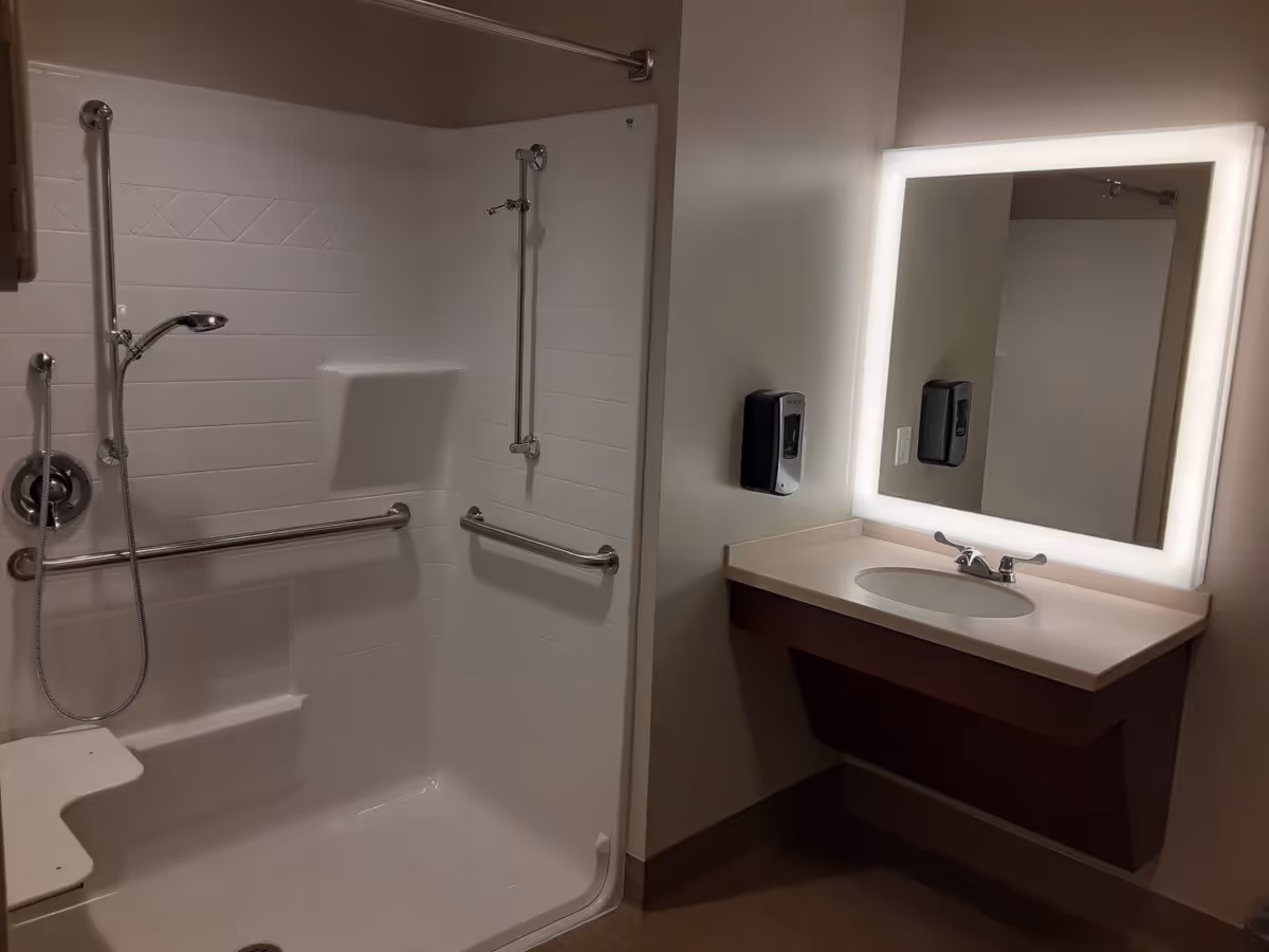 A bathroom with a walk-in shower featuring grab bars and a handheld showerhead. There is a built-in shower seat and a soap dish. Next to the shower is a sink with a countertop and a large illuminated mirror above it. A soap dispenser is mounted on the wall beside the sink.