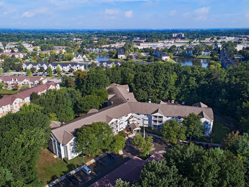 Aerial view of a senior living facility named American House Westland Hunter surrounded by trees and greenery, with multiple buildings, parking areas, and a pond visible in the background under a partly cloudy sky.