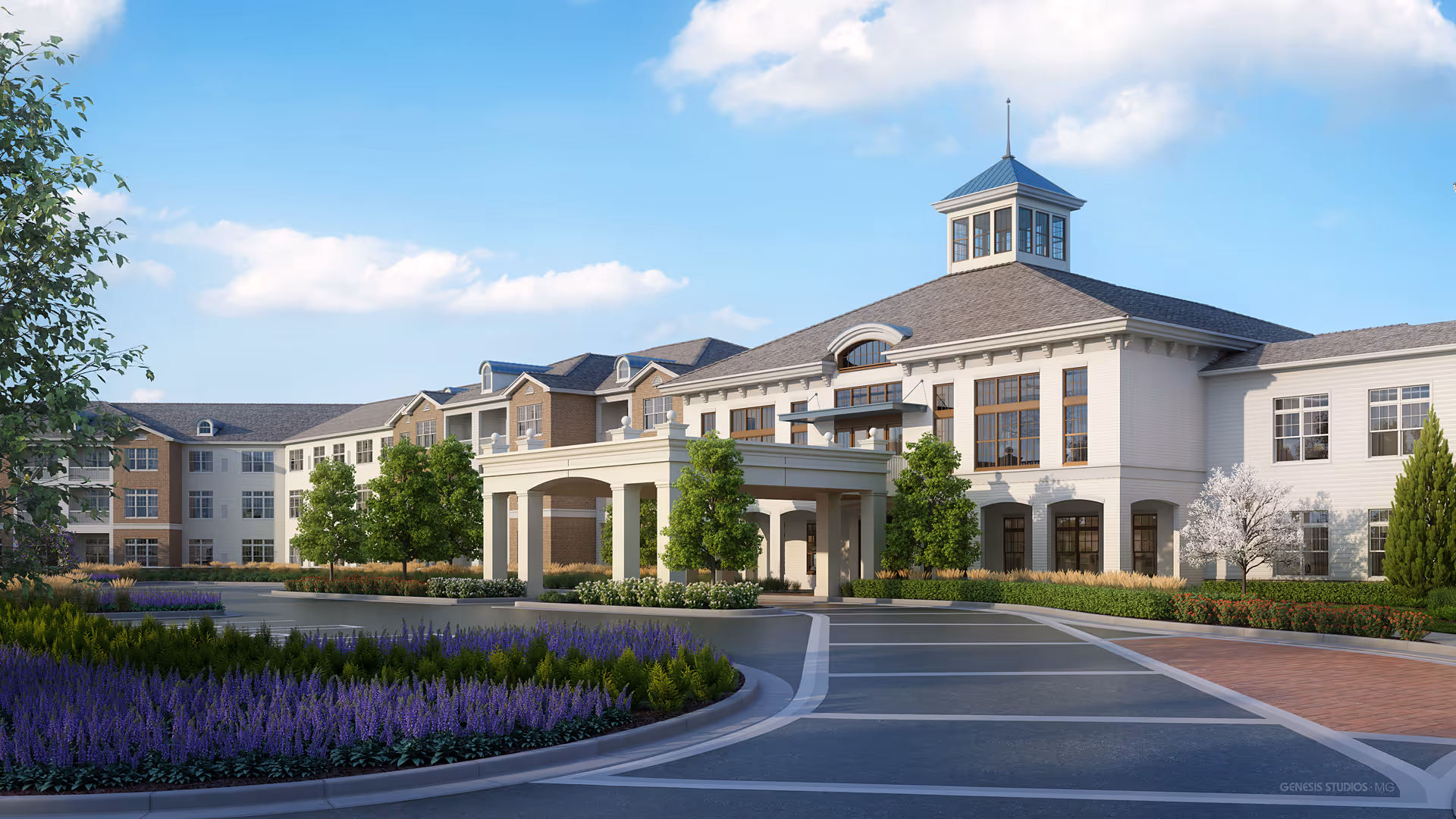 Front exterior view of a large, elegant senior living facility building with a covered entrance, multiple windows, and a cupola on the roof. The driveway is surrounded by landscaped gardens with purple flowers, green shrubs, and trees under a partly cloudy blue sky.