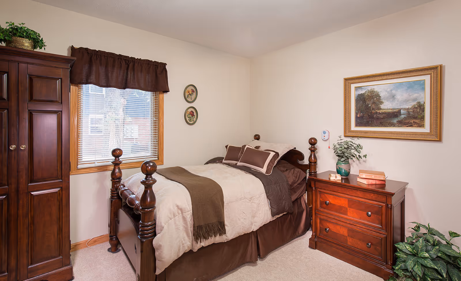 A neatly furnished bedroom with a wooden four-poster bed, matching dresser and armoire, a window with blinds, and framed wall art.