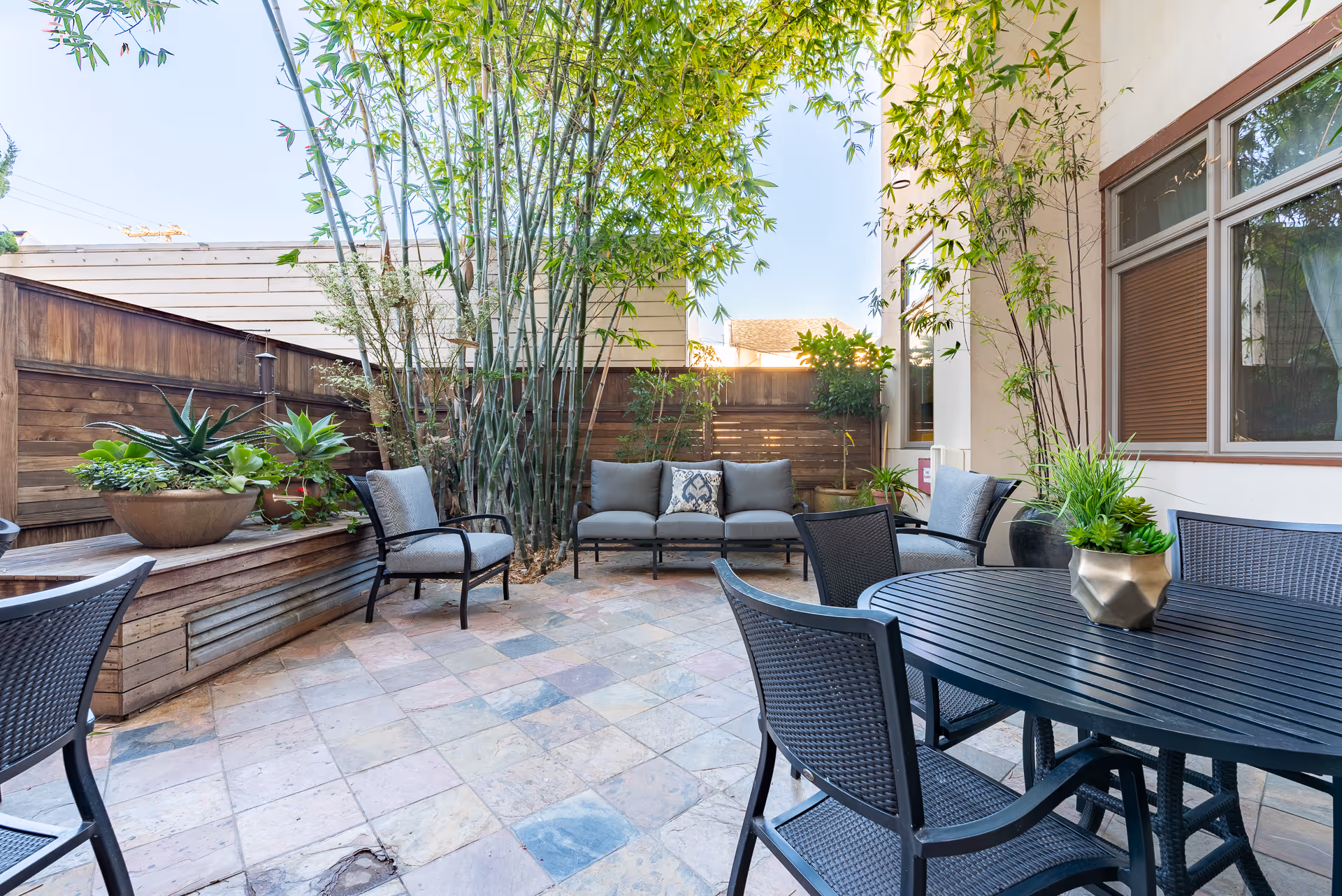 Private sunlit patio with a round dining table, chairs, cushioned seating and potted plants against a bamboo-lined wooden fence.