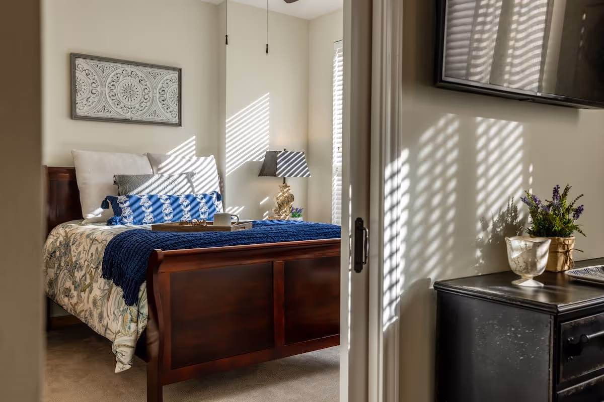 A cozy bedroom with a wooden bed frame, a bed made with patterned bedding, multiple pillows, and a blue throw blanket. A bedside table with a decorative lamp is next to the bed. Sunlight filters through window blinds, casting shadows on the walls and furniture. A flat-screen TV is mounted on the wall above a black dresser with decorative items including a small plant and a bowl.