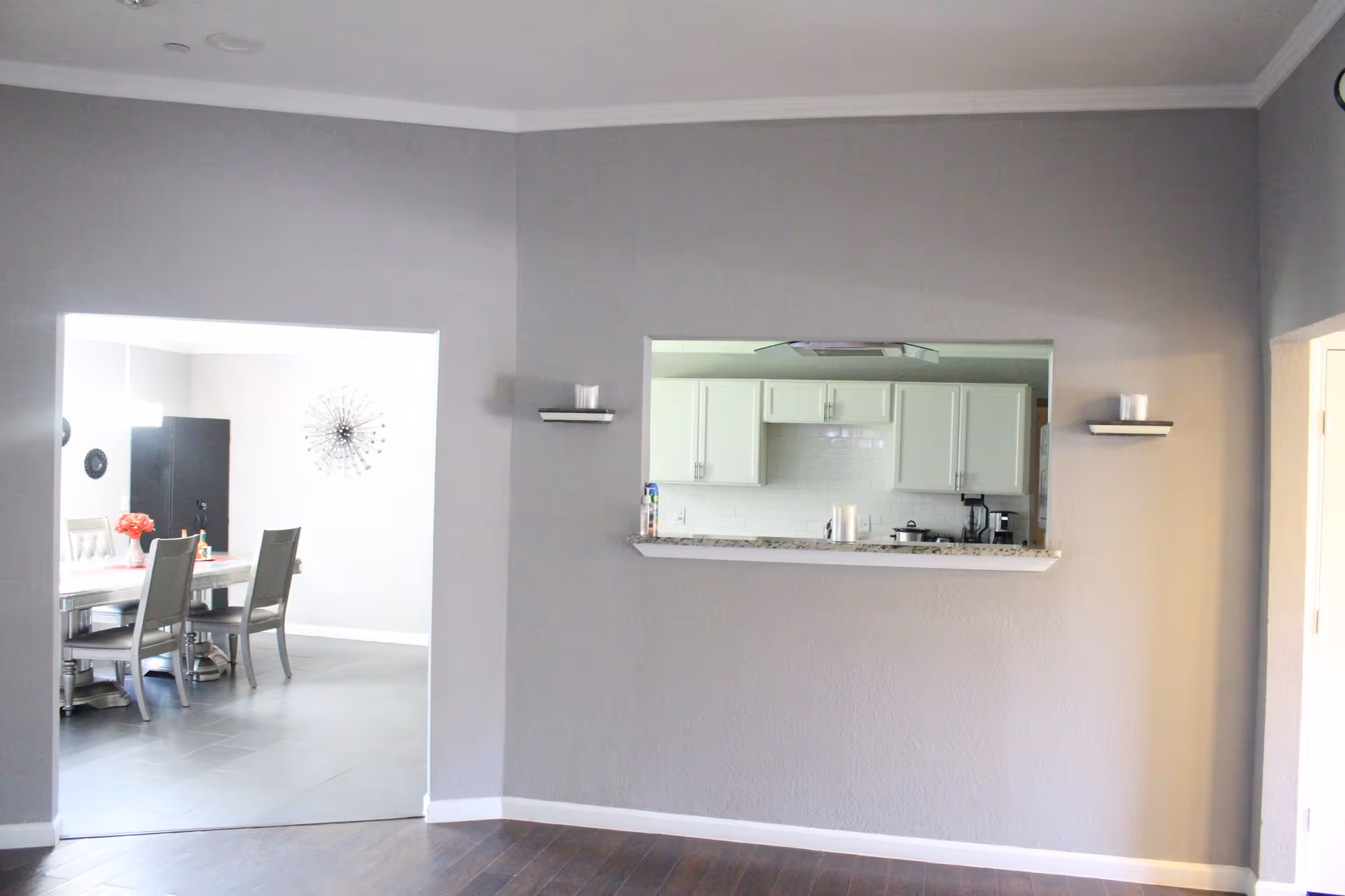 Interior view showing a gray wall with a pass-through window into a kitchen with white cabinets and a granite countertop. To the left, there is an open doorway leading to a dining area with a table, chairs, and wall decor. The floor is dark wood in the foreground and gray tile in the dining area.