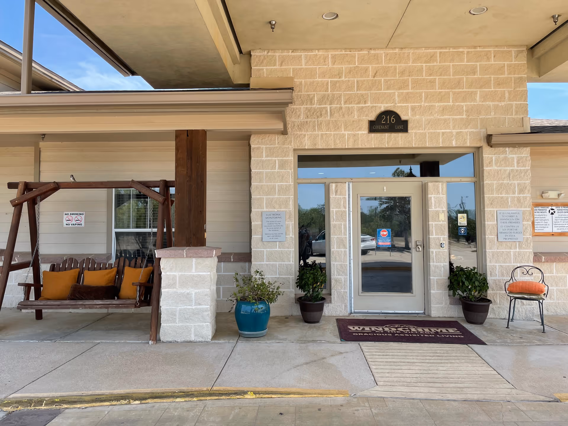 Entrance to Windchime at the Village Assisted Living facility with a glass door, two potted plants on either side, a wooden swing bench with orange cushions on the left, and a single chair with an orange cushion on the right. The building exterior is light-colored brick and siding with a sign above the door reading '216 Covenant Lane'.