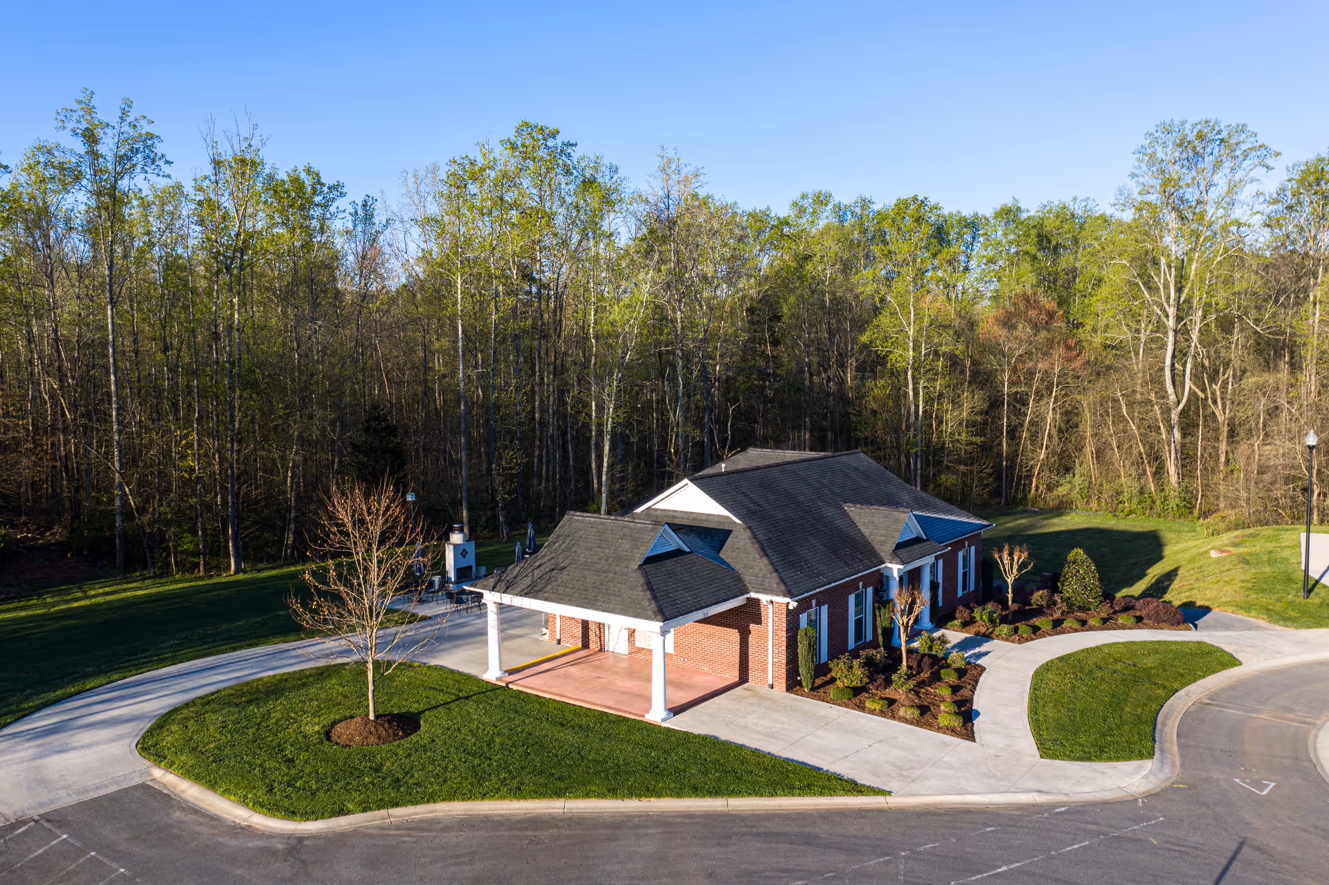 A single-story brick building with a covered entrance, surrounded by landscaped greenery and a curved driveway, set against a backdrop of tall trees under a clear blue sky.