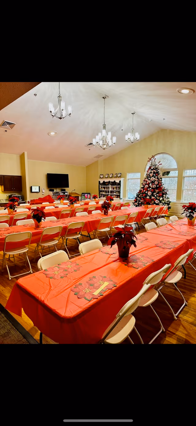 A festive dining room decorated for the holidays with long tables covered in red tablecloths and poinsettia centerpieces. There is a large decorated Christmas tree near the windows, and the room is well-lit with chandeliers hanging from the ceiling. Chairs are arranged neatly around the tables, and a television and bookshelf are visible in the background.