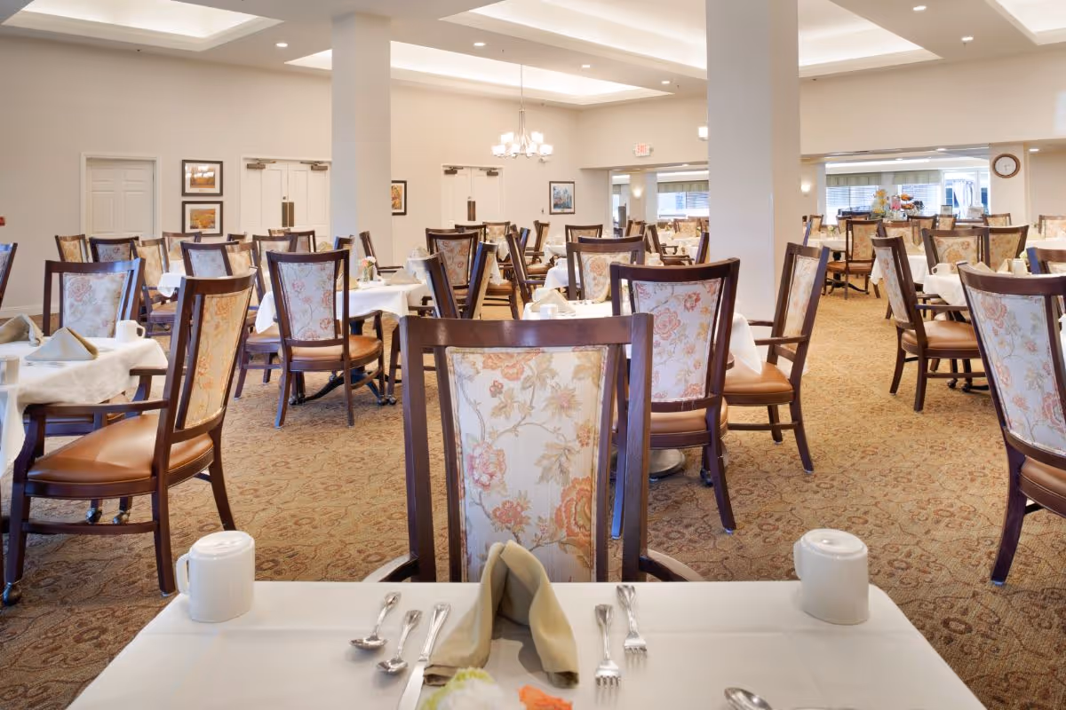 A spacious dining room with multiple tables covered in white tablecloths, each set with silverware, napkins, and cups. The chairs have floral upholstery and wooden frames. The room is well-lit with ceiling lights and chandeliers, and there are beige walls and carpeted floors.