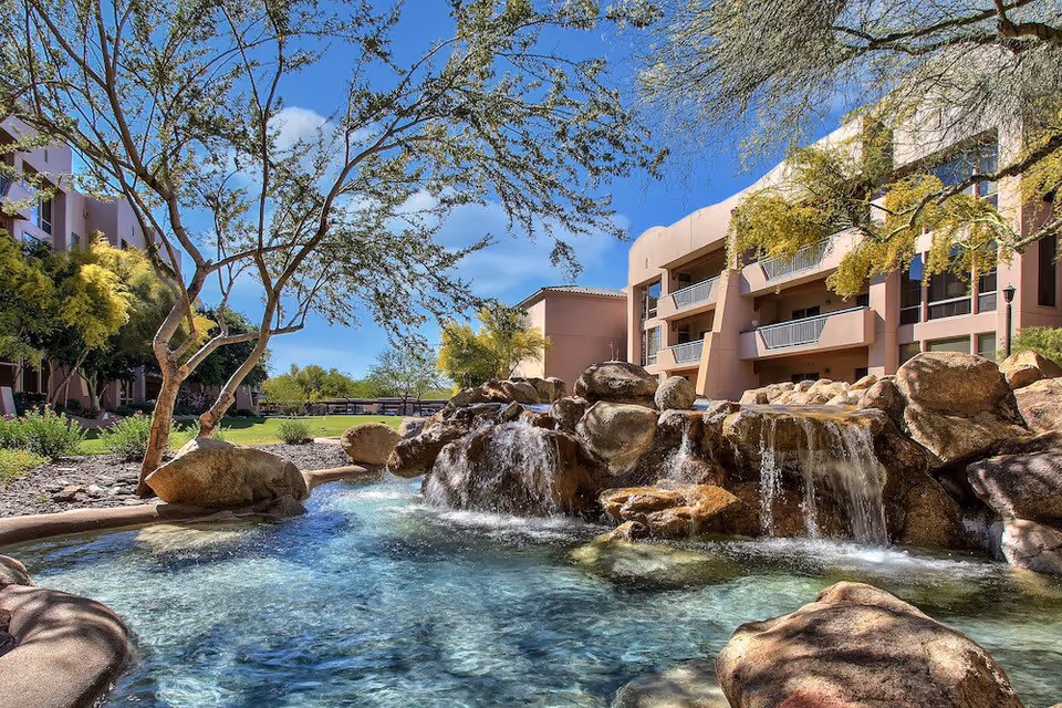 Outdoor view of a senior living facility with a clear blue sky, trees, and a rock waterfall feature flowing into a small pool. The building has balconies overlooking the water feature and landscaped grounds with green grass and shrubs.