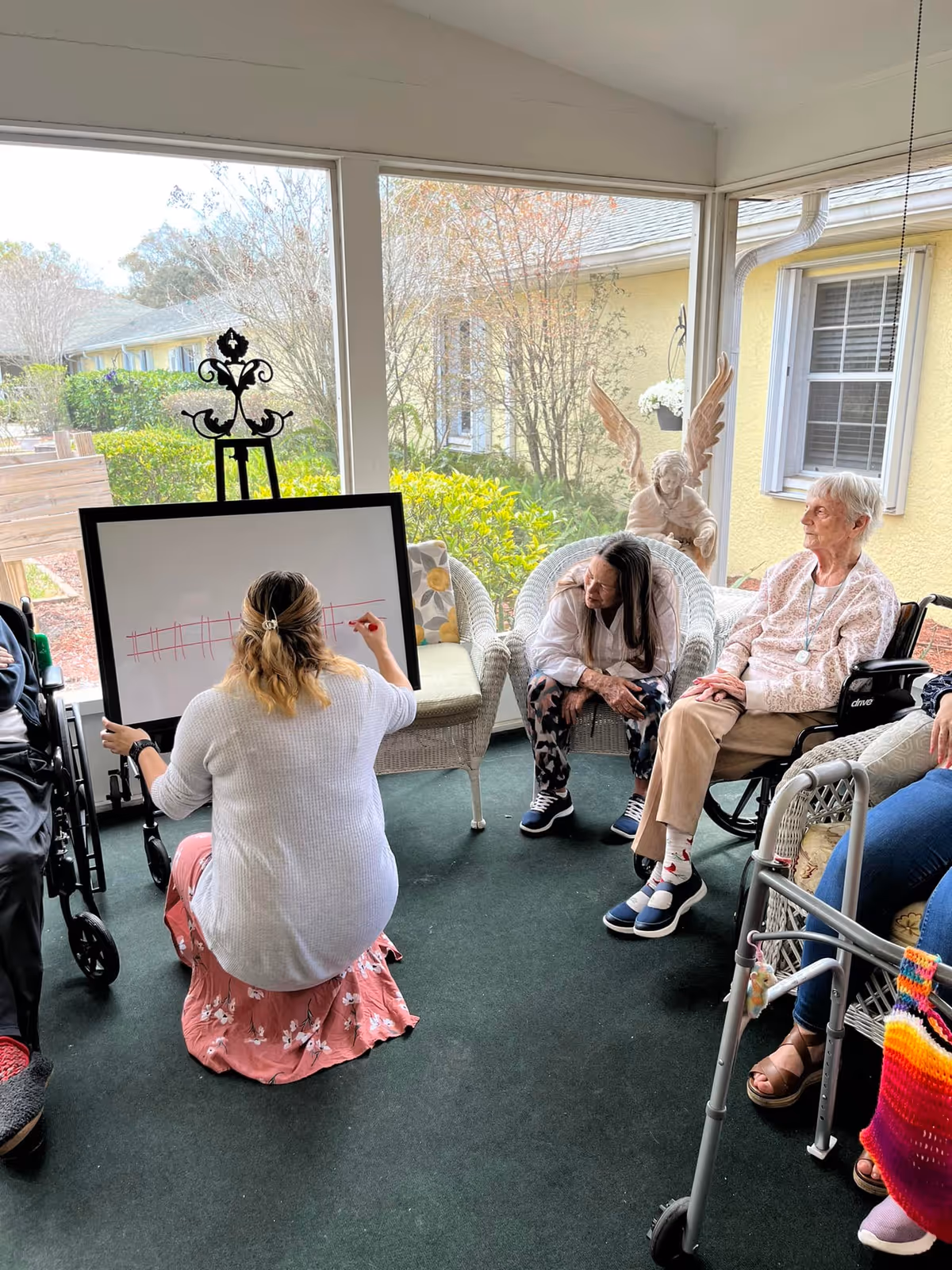 A group of elderly people sitting in a sunroom or enclosed porch area, watching a woman who is kneeling on the floor and drawing on a whiteboard. The room has large windows showing greenery outside, and there is a decorative angel statue in the background.