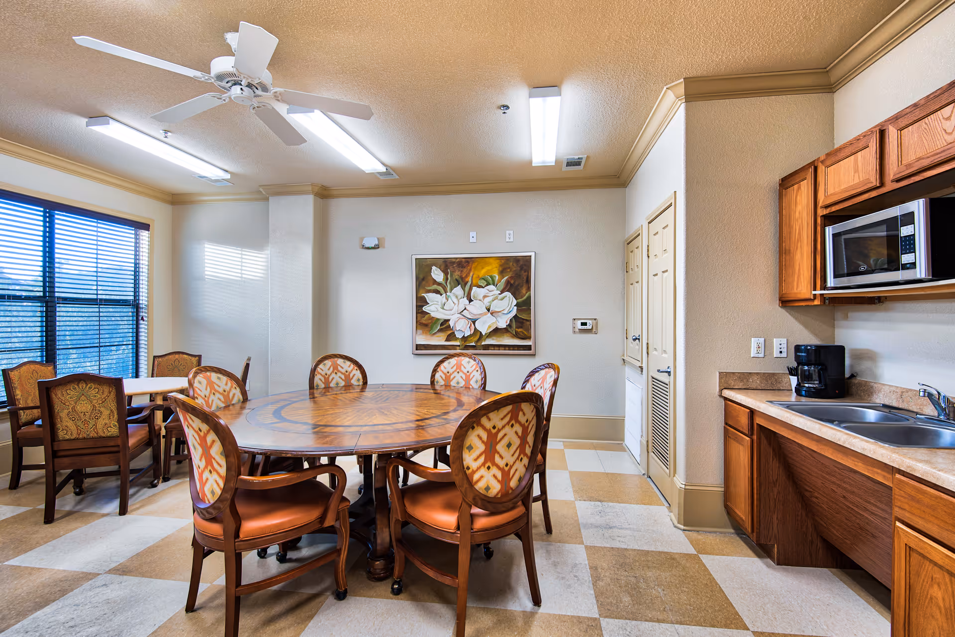 A dining area in a senior living facility with a large round wooden table surrounded by six upholstered chairs. To the right, there is a kitchenette with wooden cabinets, a microwave, a coffee maker, and a sink. The room has a ceiling fan, fluorescent lighting, a large window with blinds, and a painting of white flowers on the wall.