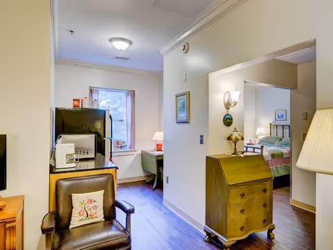 Interior view of a cozy retirement unit showing a sitting area with an armchair, a small kitchenette area with appliances, and a glimpse into a bedroom.