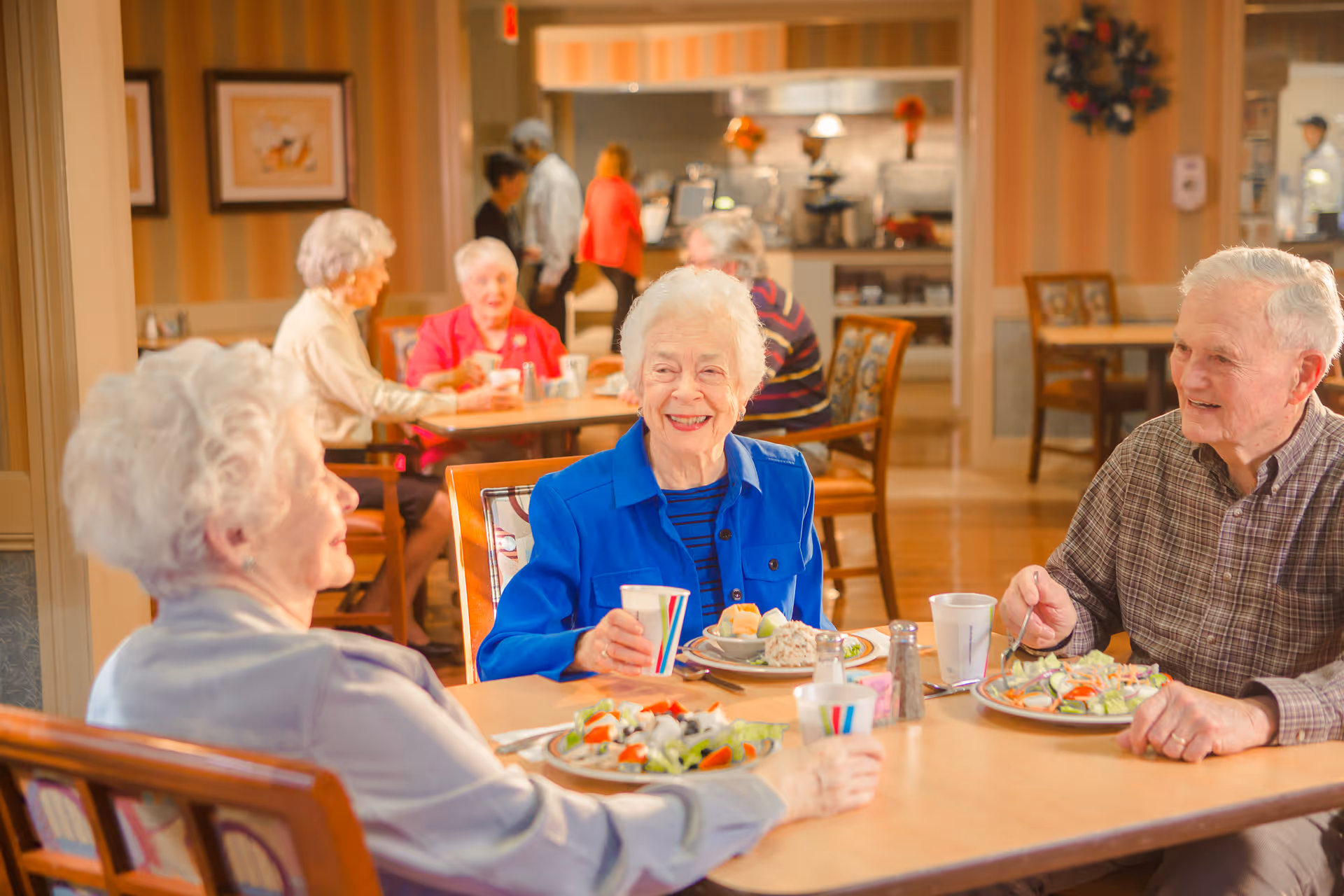 Three elderly people sitting around a table in a dining area, enjoying a meal and drinks, with other seniors and staff visible in the background in a warm, well-lit room.