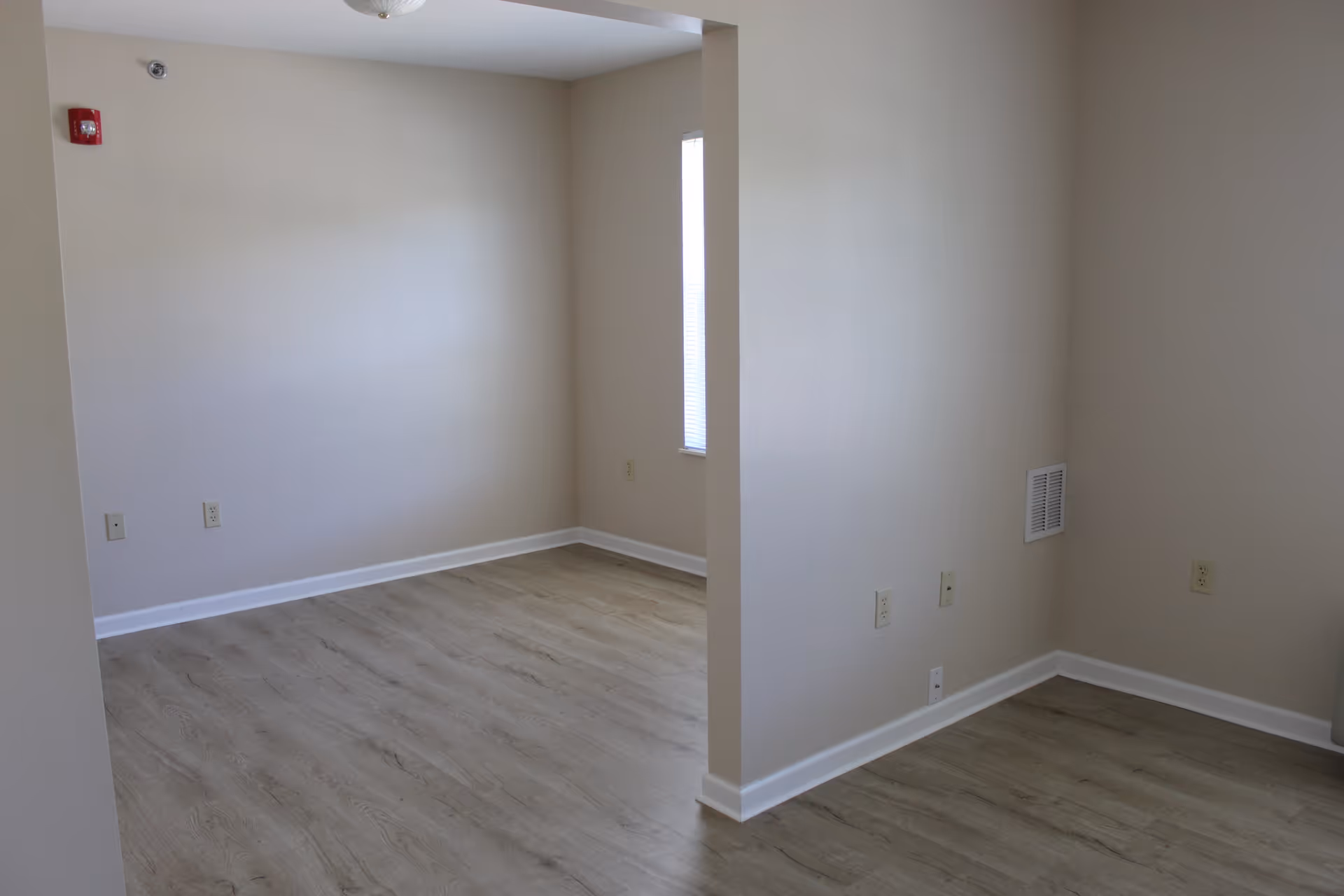 Empty interior room with light beige walls and wood-patterned flooring, featuring a small window with blinds and multiple electrical outlets along the walls.